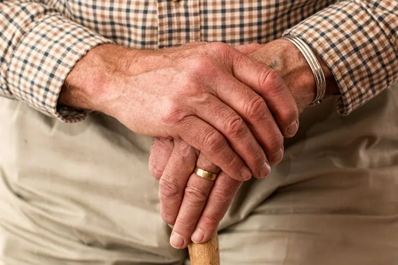 An elderly man holding a walking stick in a close-up view, highlighting his aged hands.