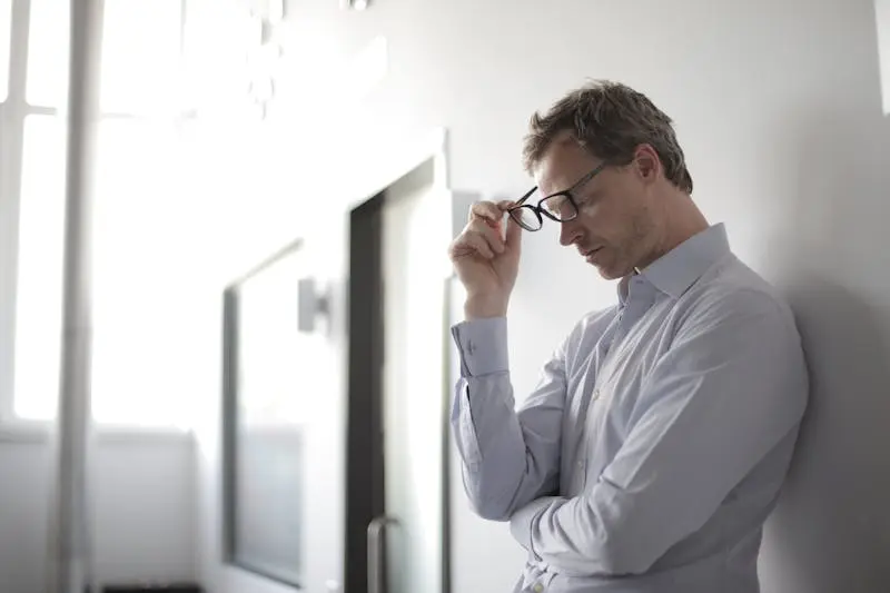 A man with glasses stands against a wall, appearing stressed.