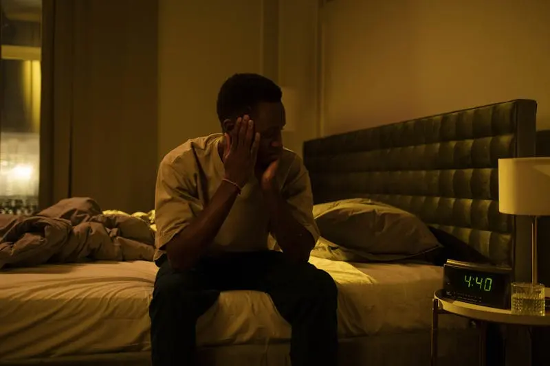 A man seated on a bed, surrounded by darkness, looking concerned.